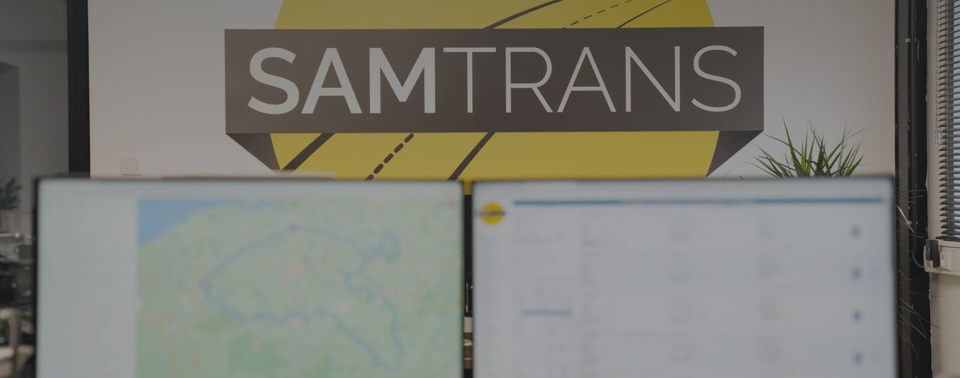 Computerschermen met maps en planningsysteem van distributiebedrijf SamTrans met logo en kantoorruimte op de achtergrond.